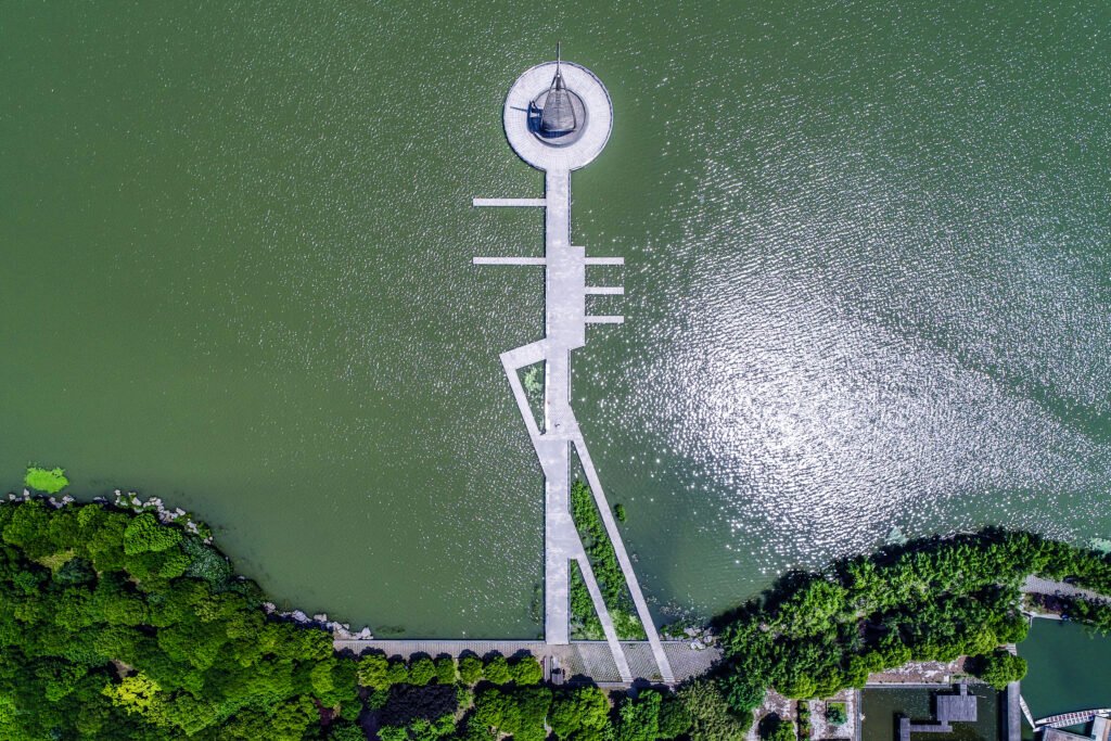 Aerial top-down view of a geometric modern concrete pier and circular observation deck extending into a green lake surrounded by lush trees.