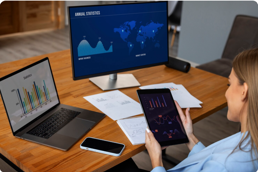 A professional woman analyzing comprehensive business data across a laptop, a large desktop monitor, and a tablet, all displaying "Annual Statistics" and global market charts.
